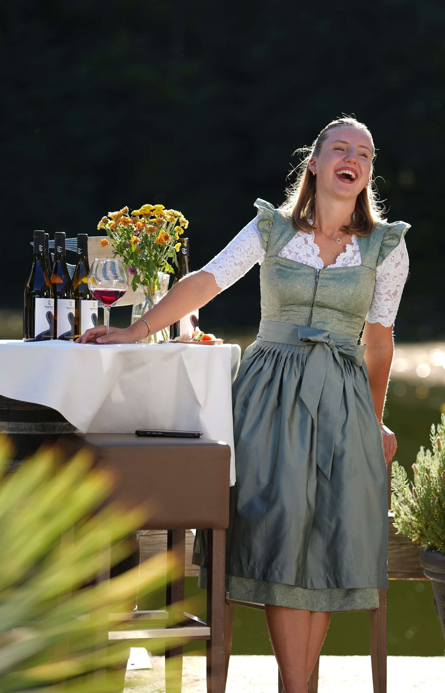 Junge Frau im grünen Dirndl lacht am Tisch mit Weinflaschen und Blumen